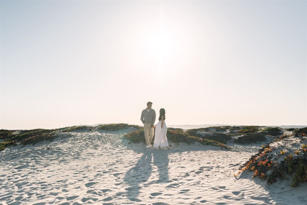 A couple stands on the sunlit sand dunes at Coronado Beach during their engagement session, beautifully captured by a San Diego engagement photographer.